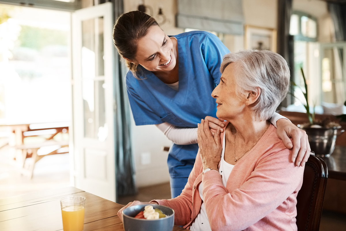 A caregiver in blue scrubs warmly smiles at an elderly woman, who sits at a table with a bowl of fruit.