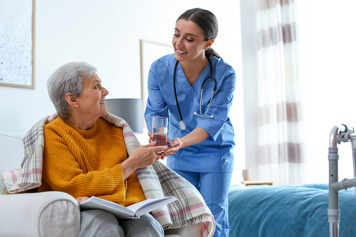 A nurse in blue scrubs, smiling, hands a glass of water to an elderly woman in a mustard sweater.